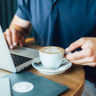 Foto: Eine Person hält eine Tasse Kaffee und sitzt vor ihrem Laptop.