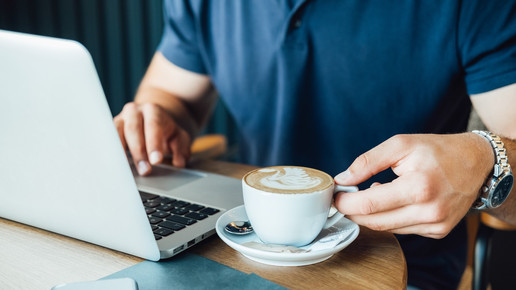 Foto: Eine Person hält eine Tasse Kaffee und sitzt vor ihrem Laptop.
