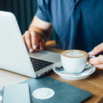 Foto: Eine Person hält eine Tasse Kaffee und sitzt vor ihrem Laptop.