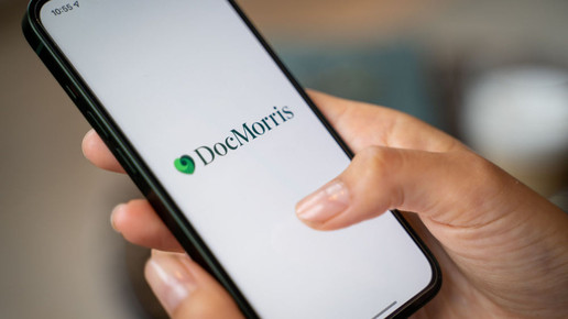 Foto: Eine Person hält ein Handy in der Hand, auf dem ein weißer Hintergrund mit zentriertem DocMorris-Logo angezeigt wird.