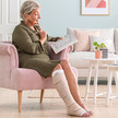 Abgebildet ist eine Dame auf einem Sessel sitzend in einem Wohnzimmer.Sie hat an ihrem rechten Unterschenkel eine SoftCompress Bandagehilfe angelegt und trägt darüber ein medizinisch adaptives Kompressionssystem in der Farbe Beige.