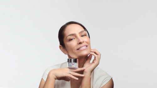 Eine Frau mit zurückgebundenem, dunklem Haar posiert lächelnd vor einem hellen, neutralen Hintergrund. Sie hält mit den Händen eine Eucerin Hyaluron-Filler Creme unter ihrem Kinn. Ihr Gesichtsausdruck ist freundlich und gepflegt.